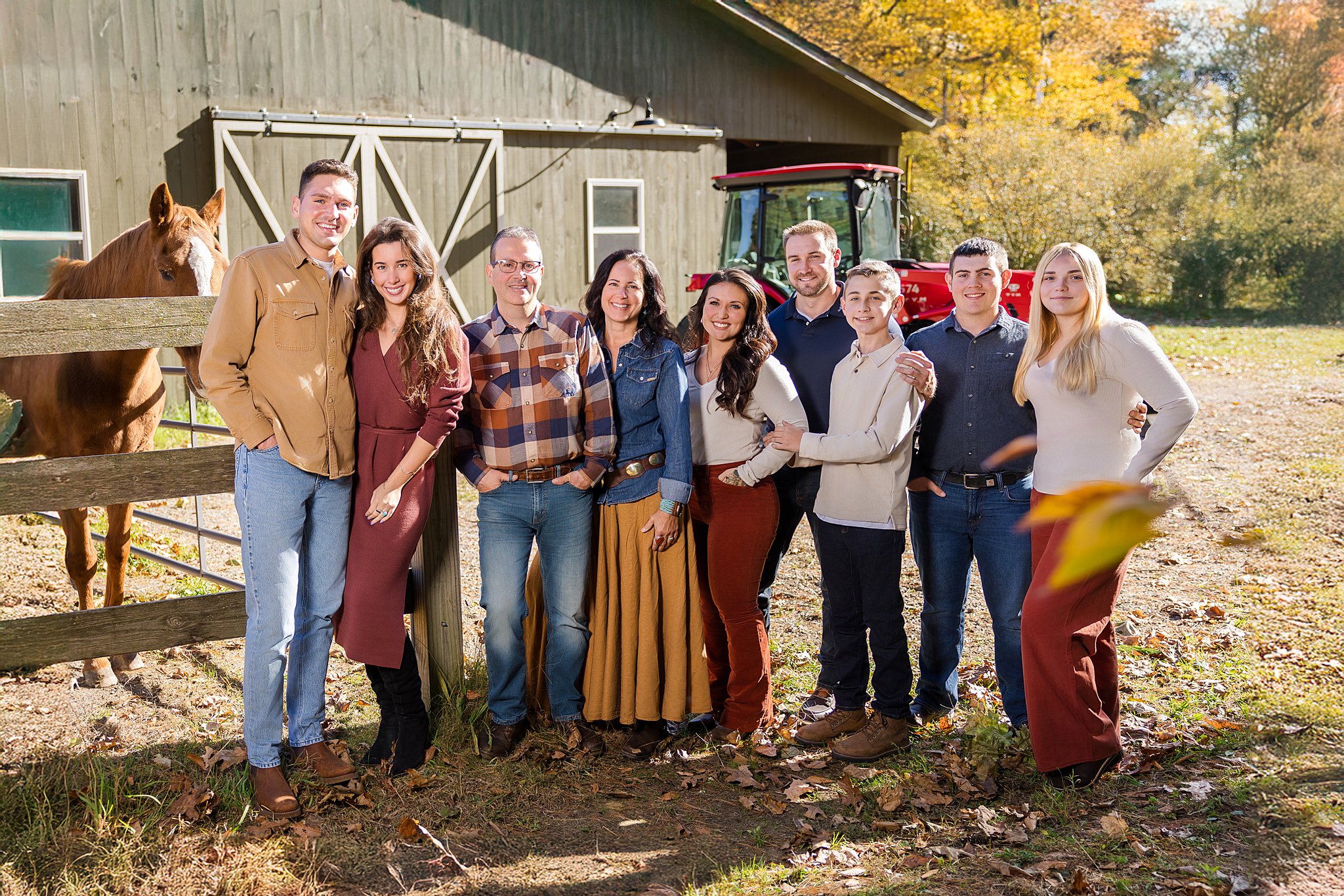 A professional portrait by a Vermont Family Lifestyle Photographer showing a large family posing outdoors near a green barn with a horse and tractor.