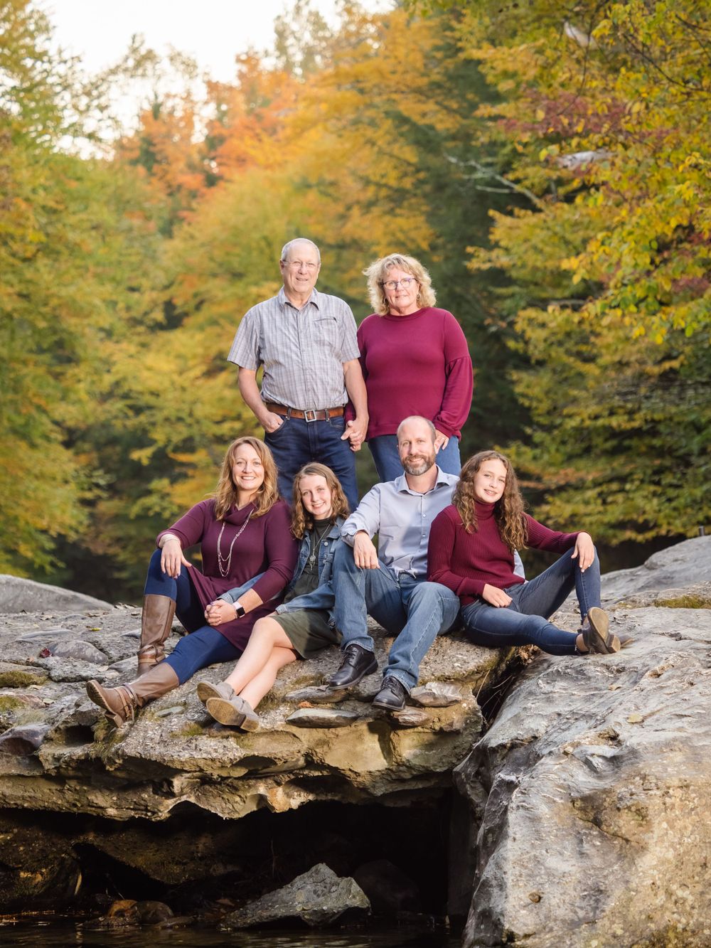 A multi-generational family sitting together on a large rock formation during a fall foliage session, captured by a professional Vermont family lifestyle photographer. The image showcases the authentic connection and vibrant autumn colors typical of high-end outdoor family portraits Vermont.