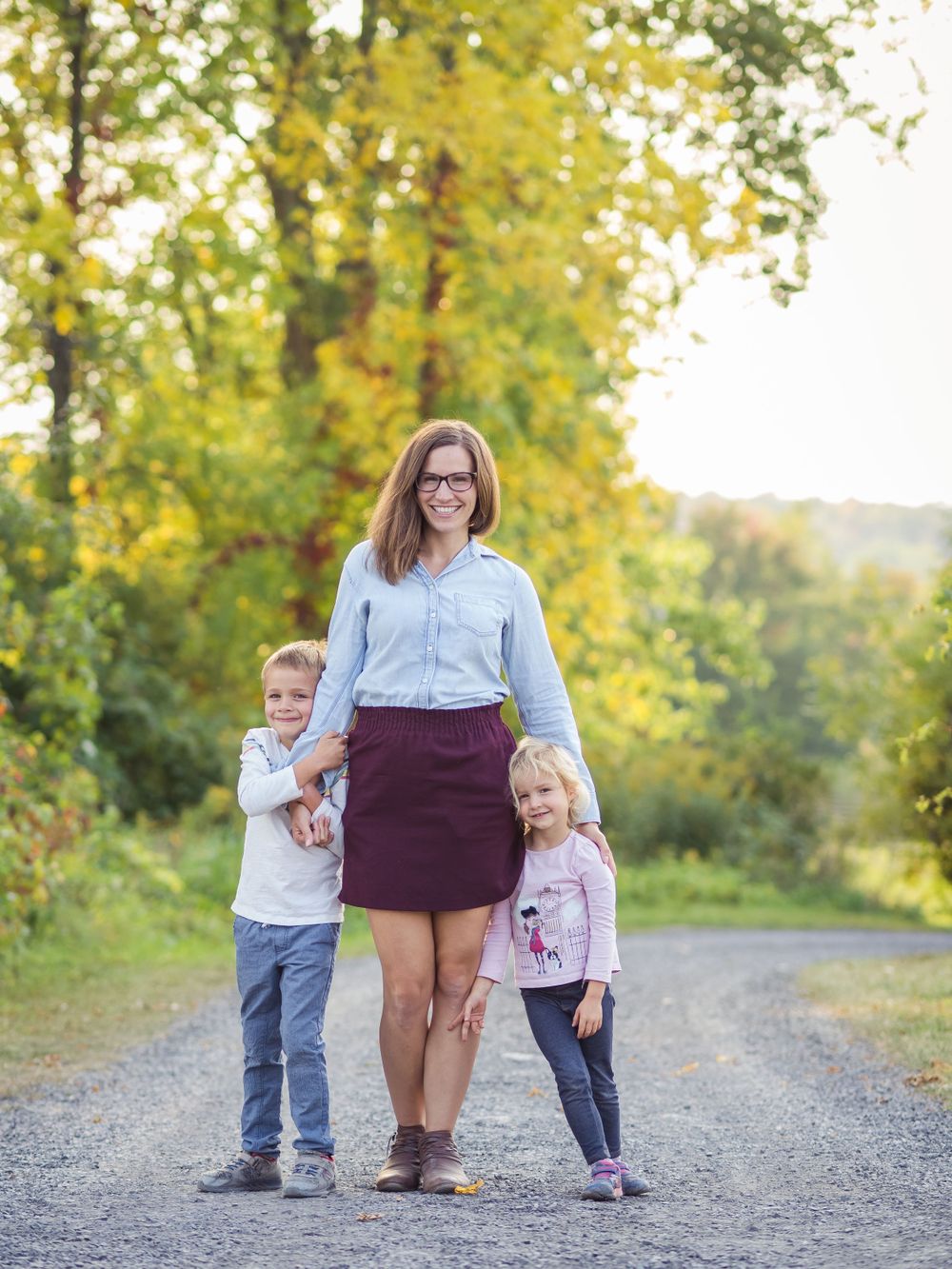 A smiling mother stands on a gravel path with her two children leaning in close for a portrait by a Vermont Family Photographer. The soft, natural light and lush green trees highlight the authentic emotional connection captured during this outdoor session