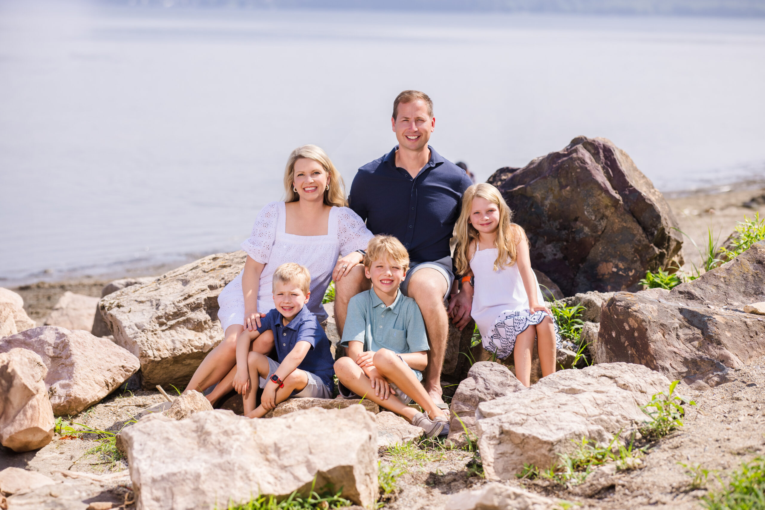 A smiling family of five—mother, father, and three children—poses together on large grey rocks by the water for a portrait from a Vermont Family Photographer. The background is a calm, hazy lake and shoreline, emphasizing the natural beauty of an outdoor session.