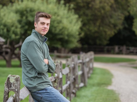 A young man leaning against a rustic wooden fence during a session with a Vermont Senior Portrait Photographer. He is wearing a green quarter-zip sweater and jeans, illustrating a classic and comfortable style for outdoor high school milestones.
