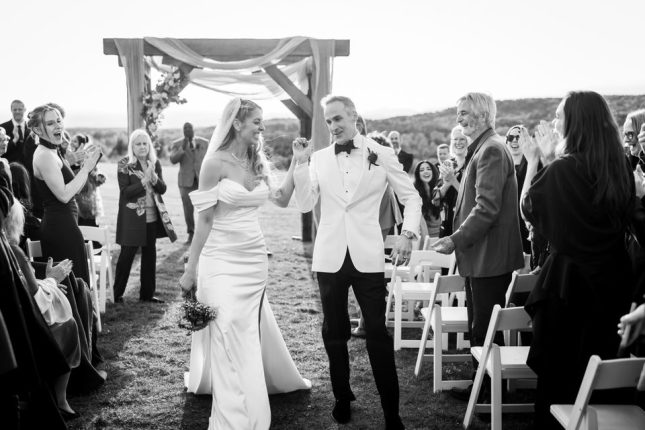 A joyful black and white image captured by a Vermont Wedding Photography professional showing a newly married couple walking down the aisle. Guests are seen clapping and cheering as the couple celebrates their first moments of marriage under an outdoor floral arbor.