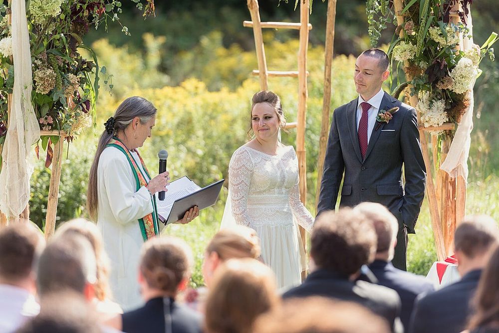 A bride and groom standing at an outdoor altar during their wedding ceremony in a Vermont field, with an officiant speaking into a microphone. This authentic moment is captured by a professional specialized in Vermont Wedding Photography.