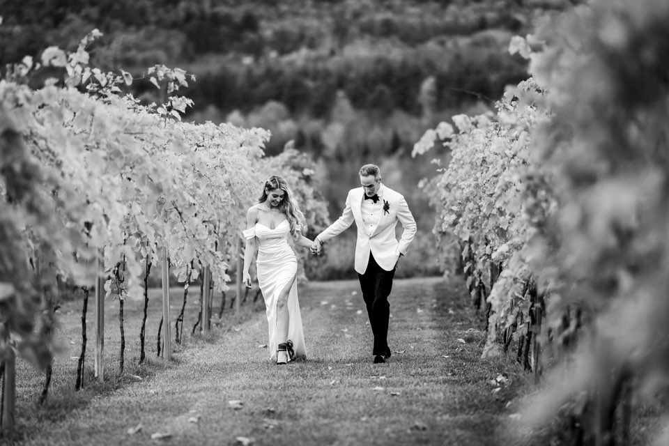 A black and white photograph by a Vermont Wedding Photography expert featuring a bride and groom holding hands while walking through a lush vineyard. The couple is dressed in elegant formal attire, showcasing the romantic and cinematic style of a Vermont wedding.