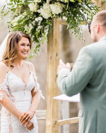 A bride smiling joyfully at her groom during their vows under a floral arbor, illustrating the emotional depth of Vermont Wedding Photography.