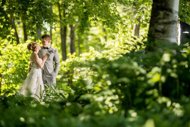 A bride in a lace gown and a groom in a gray suit sharing a candid look framed by lush green trees during a session with a Vermont wedding photographer.