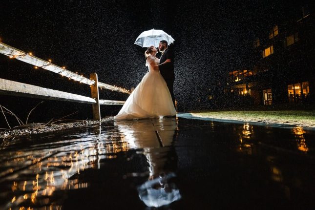A newly married bride and groom embracing under a clear umbrella during a rainy night session with a portrait photographer in Vermont.