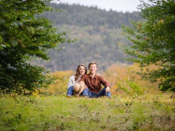 A happy couple sitting in a grassy field during an autumn engagement photos Vermont session.