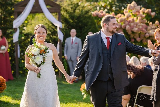 A smiling bride and groom walk down the aisle at an outdoor venue by an affordable wedding photographer near Burlington, VT.