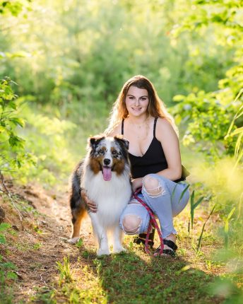 A young woman wearing a black top and ripped jeans smiles while kneeling beside her Australian Shepherd dog on a sunny woodland trail during a shoot with a professional photographer Vermont.